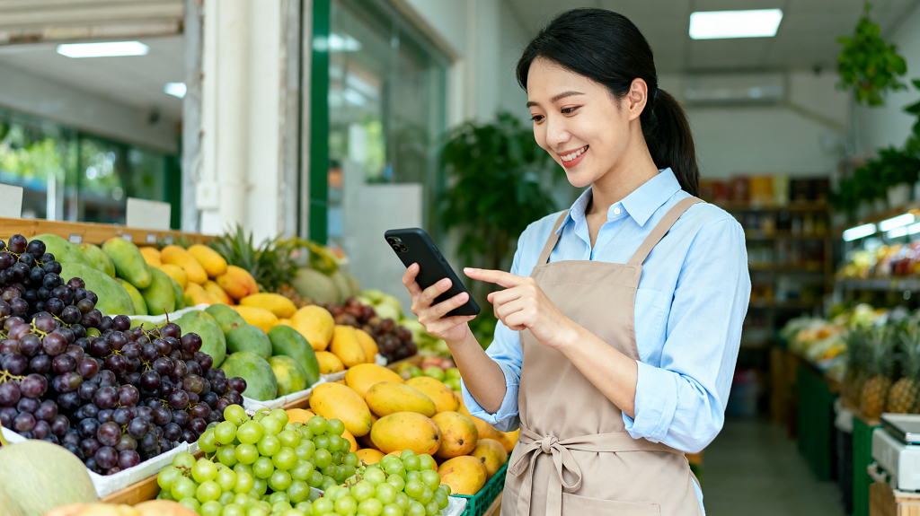 店主经营场景水果摊穿围裙女士面带微笑使用手机各色鲜果琳琅满目