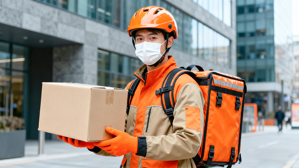 城市建筑街头外卖员橙棕制服佩戴橙色头盔口罩手套持纸箱背外卖箱