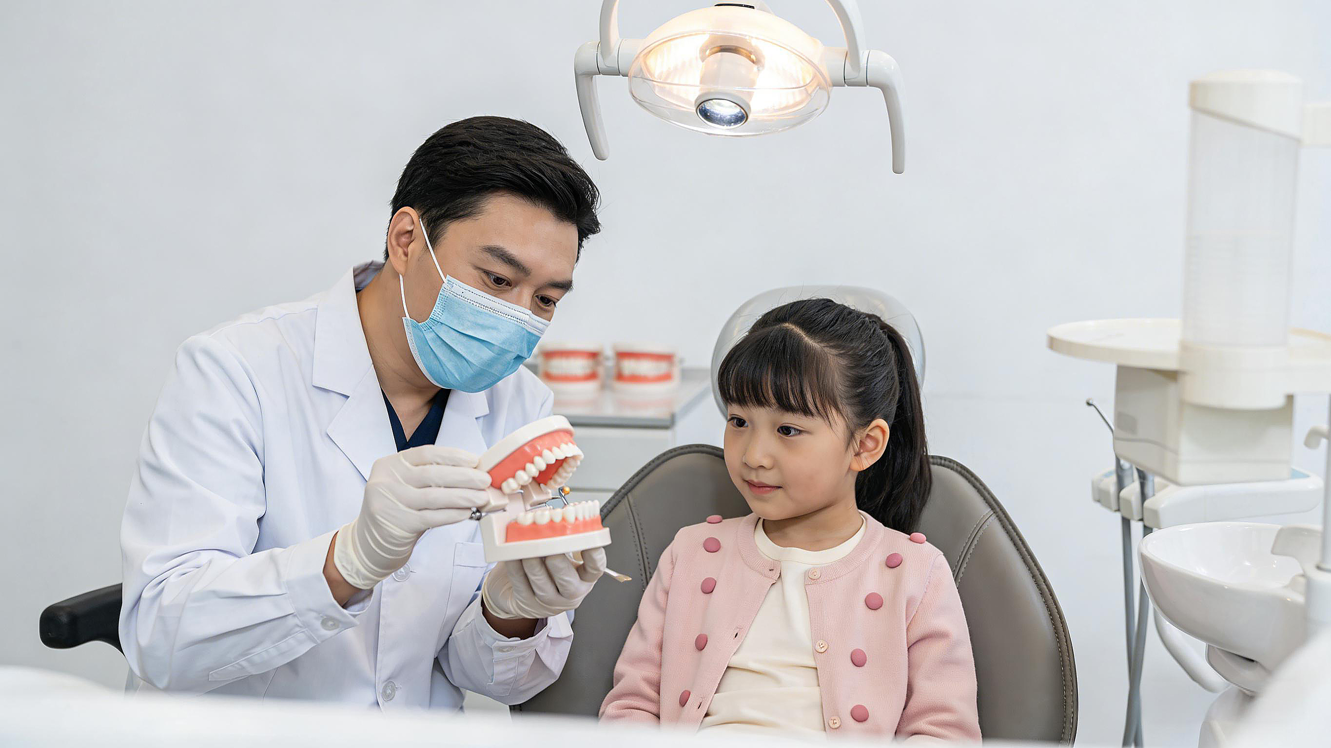 男牙医手持牙齿模型向坐在诊疗椅上的小女孩耐心讲解口腔知识