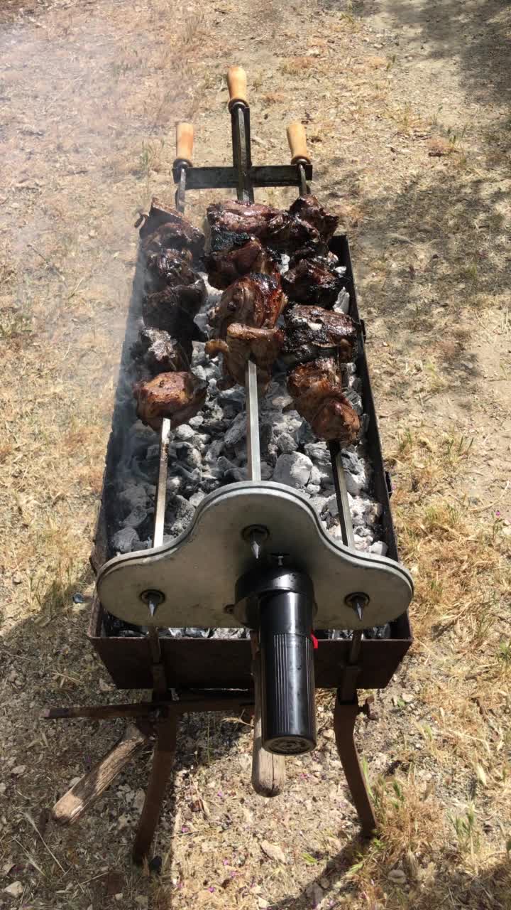 向下看，三根厚厚的、缓慢旋转的羊肉串正在烹饪，缕缕炊烟从覆盖着灰烬的木炭中升起