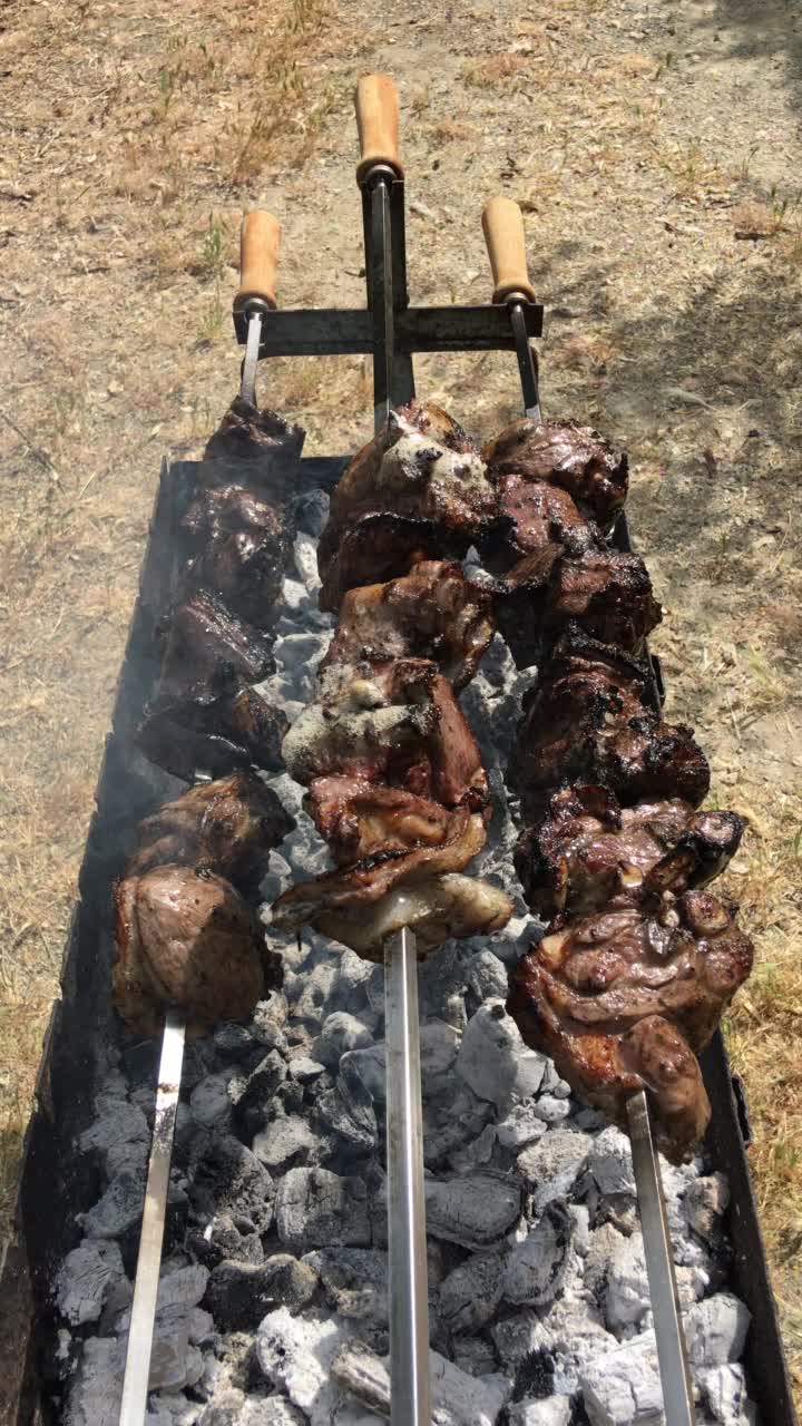 向下看三根长长的、缓慢旋转的烤肉串，上面有正在烹饪的羊肉，缕缕炊烟从覆盖着灰烬的木炭中升起