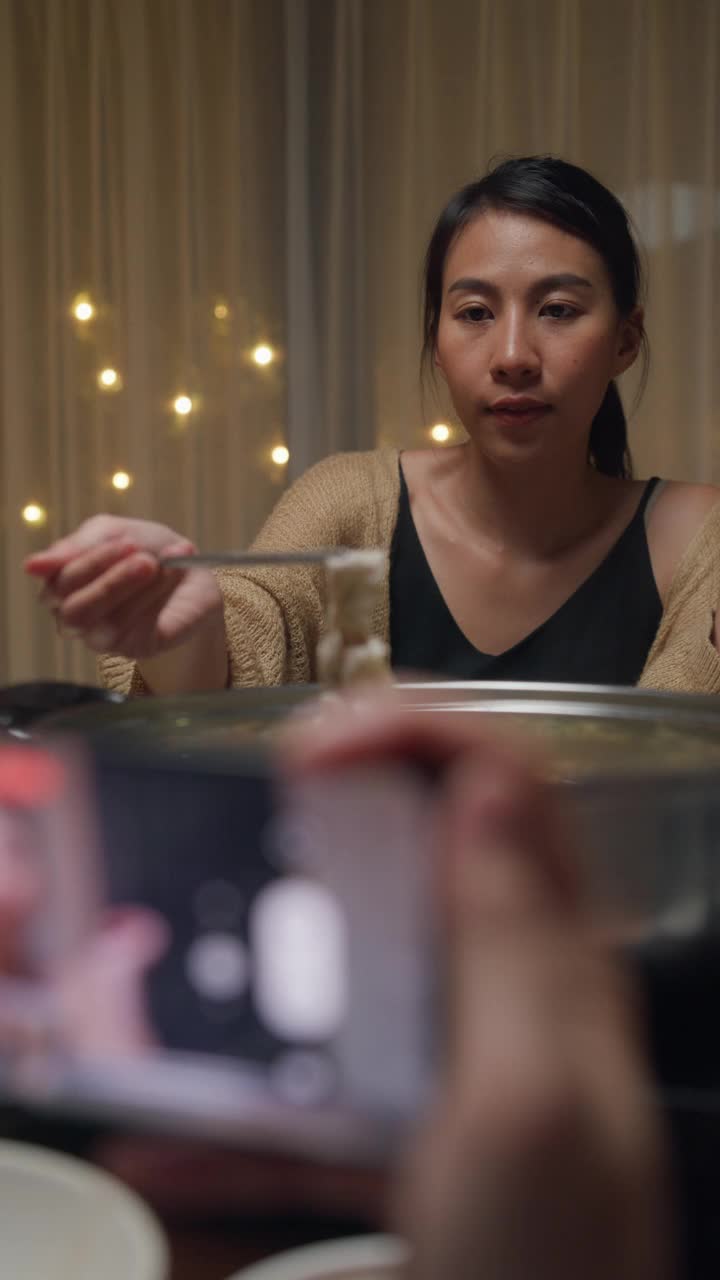 一群快乐的亚洲朋友一边吃着火锅，一边用手机拍视频，晚上在家里聚在一起吃春节团圆饭。生活方式享受美食。垂直的屏幕。