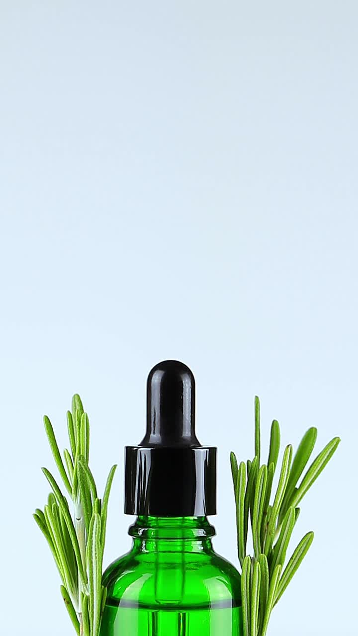 特写的女性手举滴管盖，油性的滴管落下。绿色滴瓶，内含天然化妆品油。在瓶子的每一面都放上新鲜的香草迷迭香。垂直格式视频