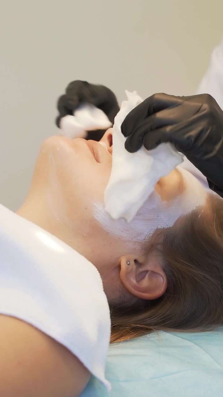 美容师从年轻女子白皙的脸上去除白色面膜。医生用白色湿海绵擦拭面部厚厚的白色物质。嫩肤、白泥、洁肤、祛痘