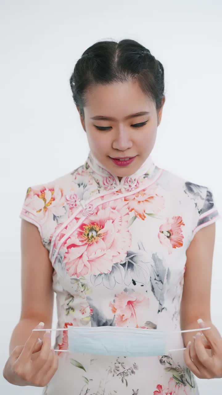中国/亚洲美丽的女子戴着护面面具，站在白色背景下，穿着旗袍，白色印花裙，看着镜头迎接新年的到来