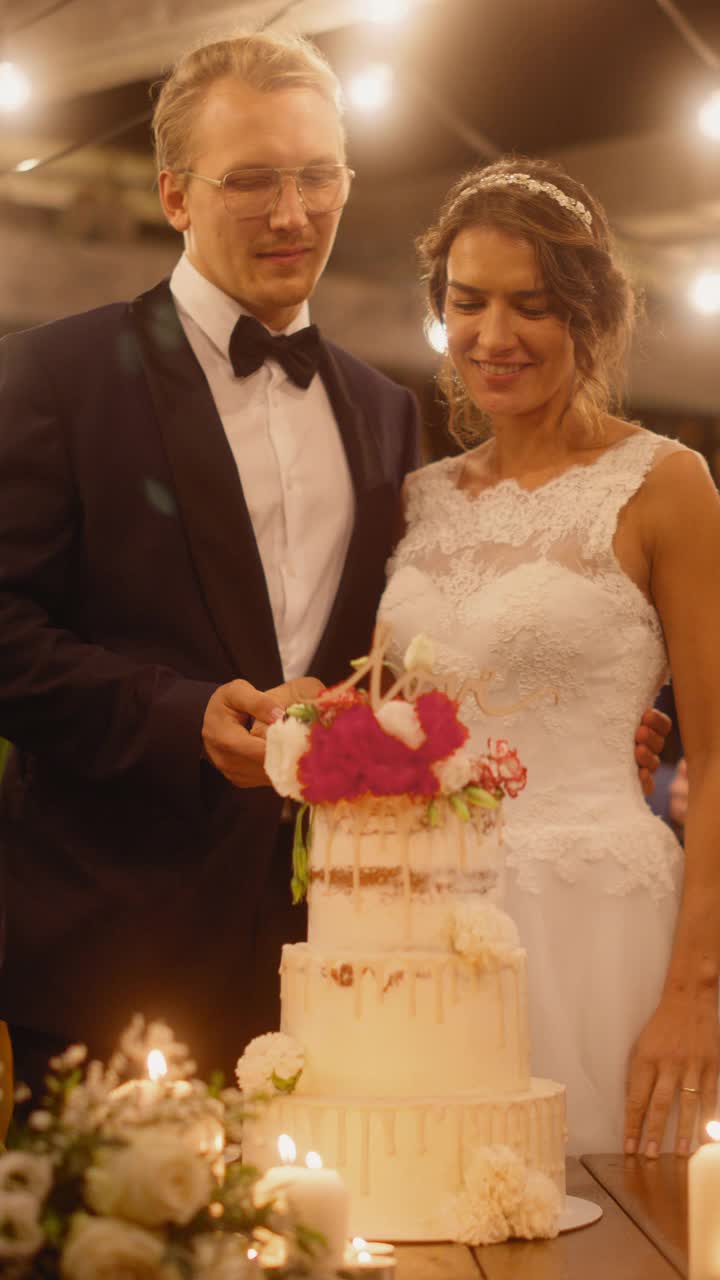 垂直画面:美丽的新娘和新郎与多民族的朋友在晚会上庆祝婚礼。已婚夫妇站在餐桌前，切婚礼蛋糕。