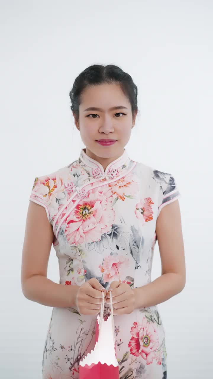 中国/亚洲妇女在春节期间打开粉红色的购物袋，在白色的背景下，穿着旗袍白色花卉图案的衣服，遇到了一个惊喜